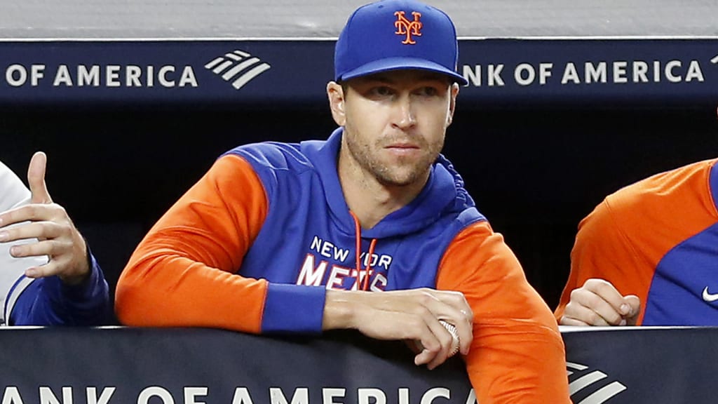 Jacob deGrom, in blue and orange Mets sweatshirt, watches a game from the dugout railing