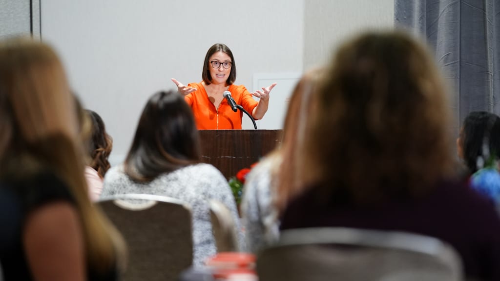 A woman stands at a podium speaking to a gathering of other women