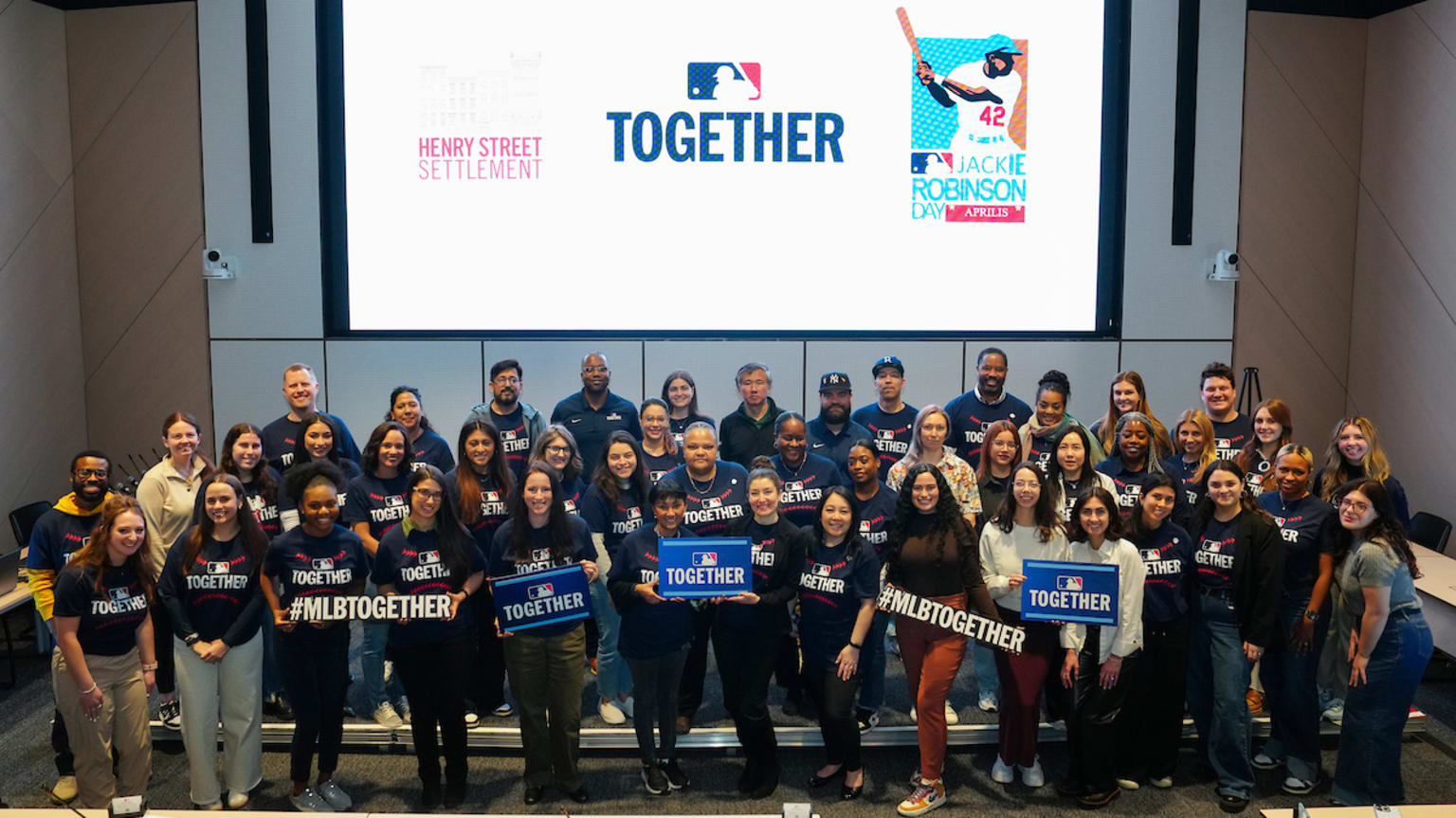 MLB employees gather for a photo under a large screen showing the logos for Henry Street Settlement, MLB Together and Jackie Robinson Day