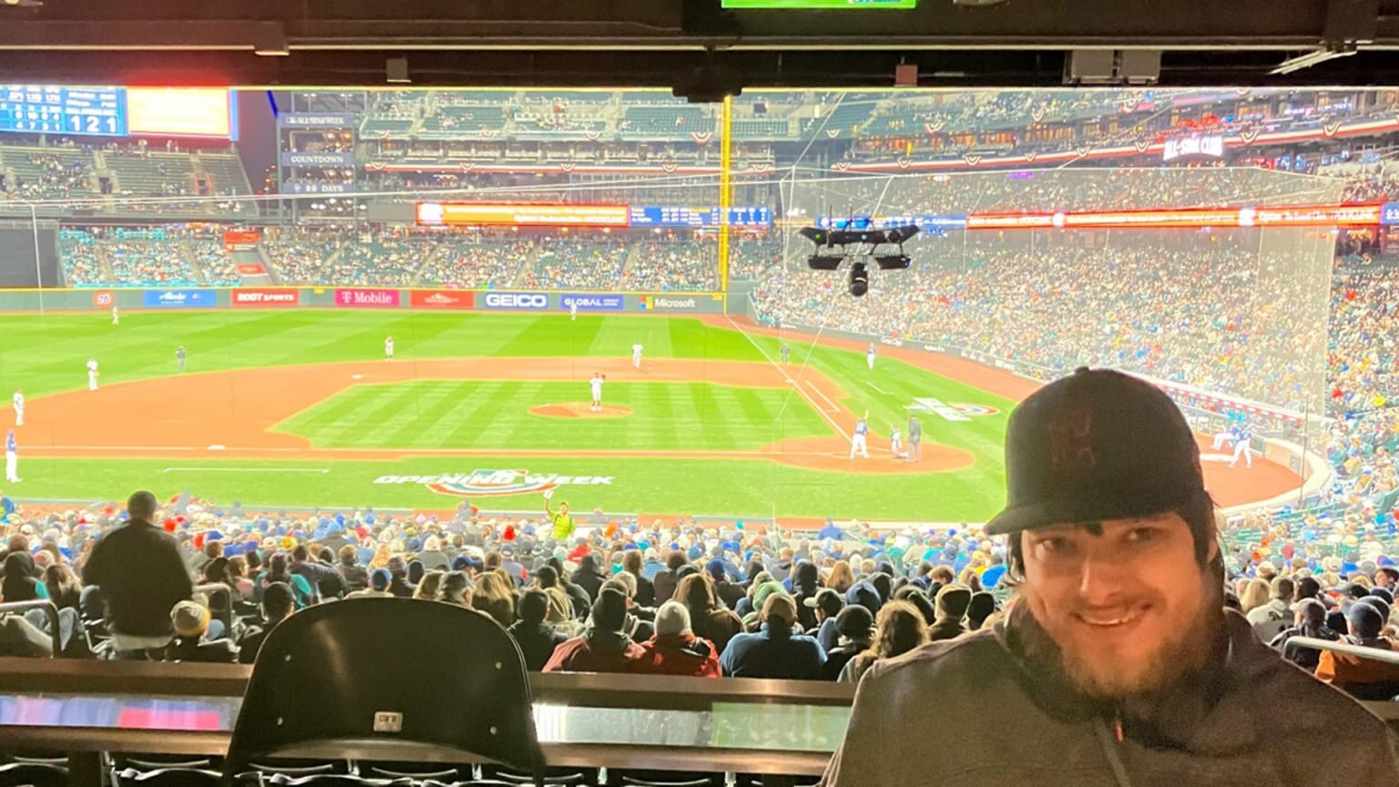 Peter Knab, wearing a Guardians cap, poses for a photo with a live baseball game in the background