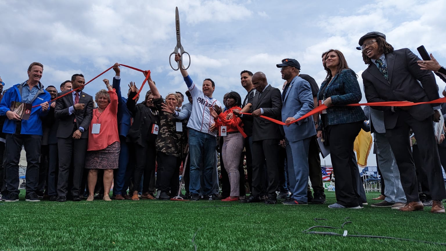 A crowd holds a red ribbon while a man in the center holds up a giant pair of scissors