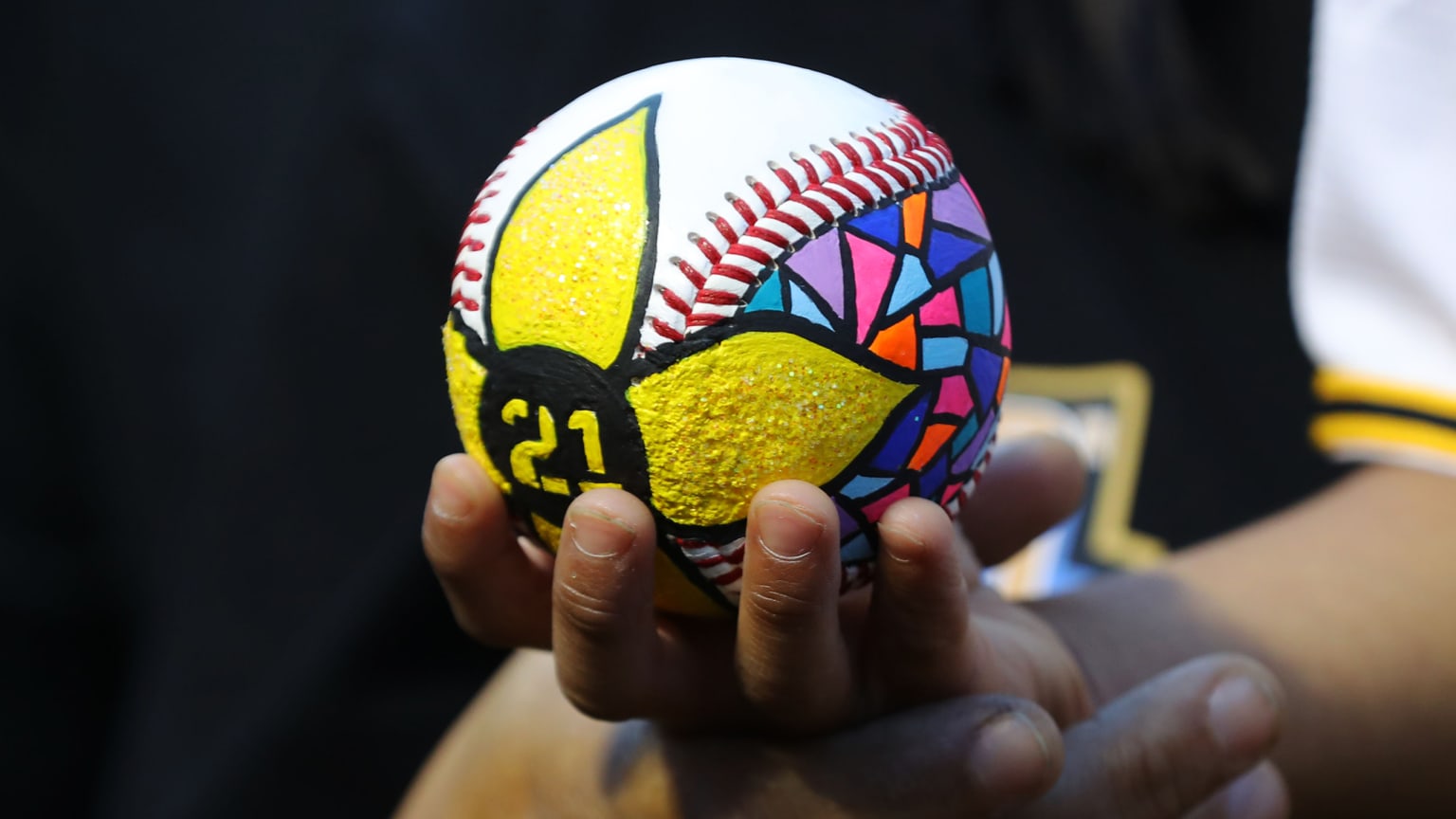 A baseball with a yellow flower painted on it and the number 21 in the center of it. To the right of the flower, multi-colored shapes resemble stained glass