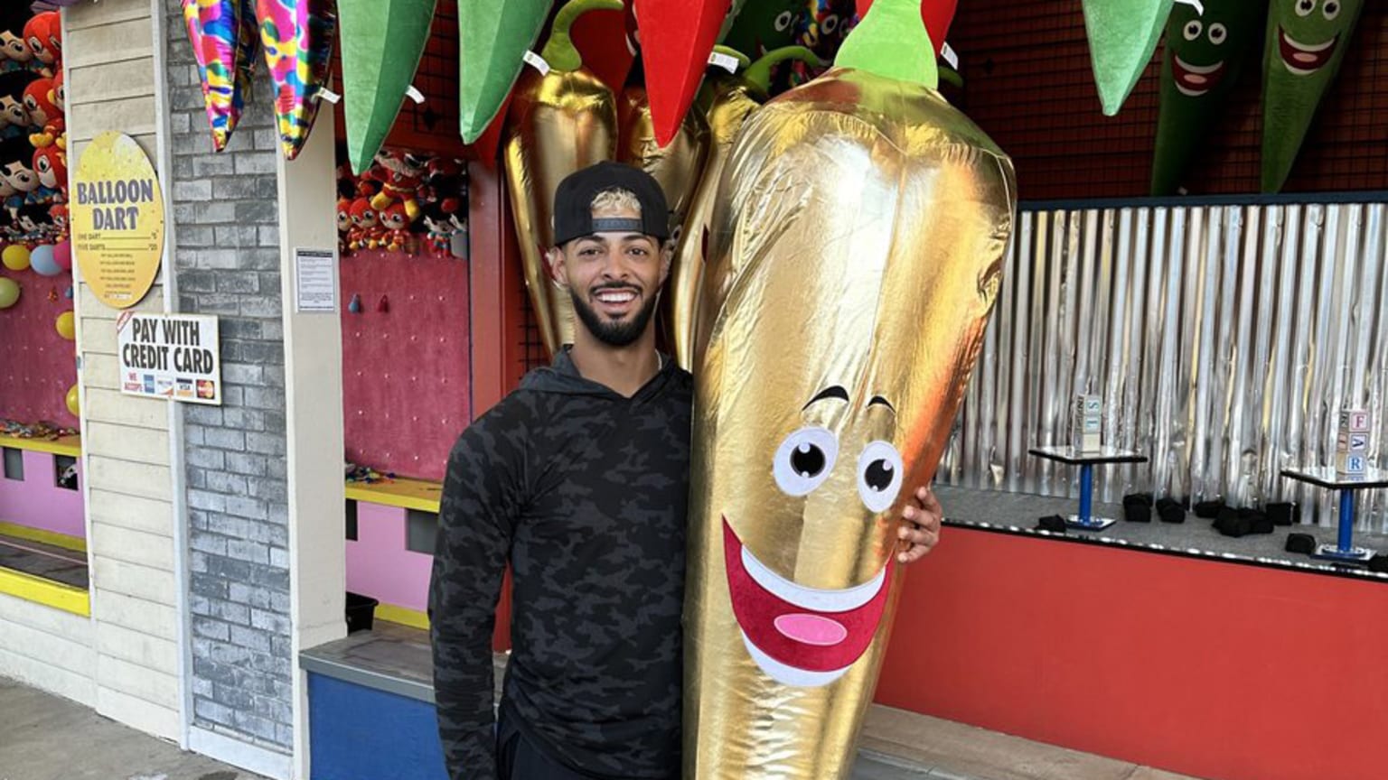 A man in a black shirt and black backwards baseball cap holds a shiny gold jalapeno as tall as he is