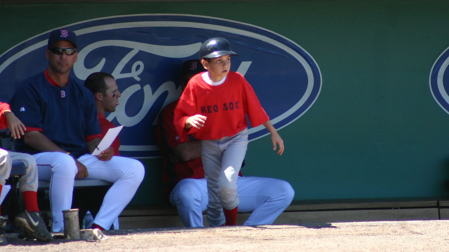 A young boy in a red T-shirt steps out of a shaded dugout into the sunshine