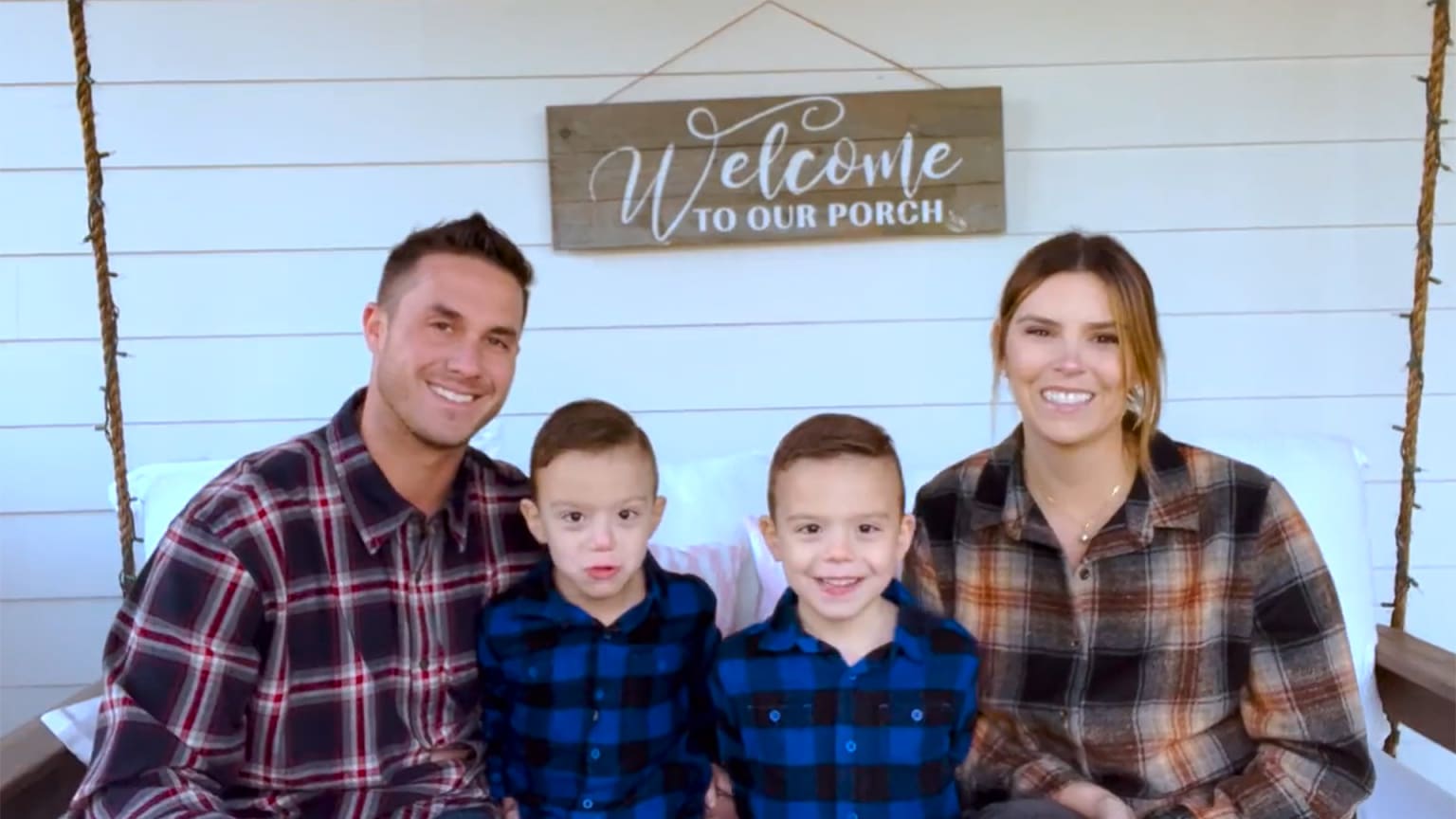 Two parents in flannel shirts sit on a porch swing with twin boys, also in flannel, between them
