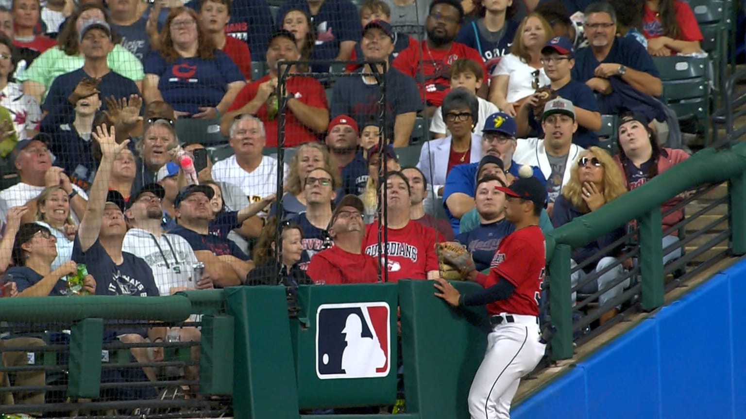 A player in a red jersey stands next to the crowd in foul territory as a baseball bounces off his head