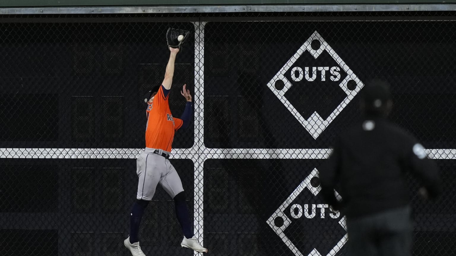 A player in an orange jersey leaps to catch a ball against a scoreboard fence