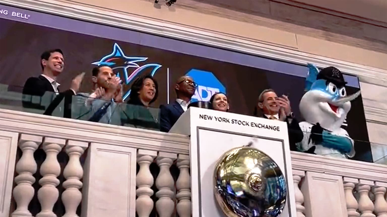 A view looking up at Kim Ng and Caroline O'Connor with four men and Marlins mascot Billy the Marlin above the bell of the New York Stock Exchange