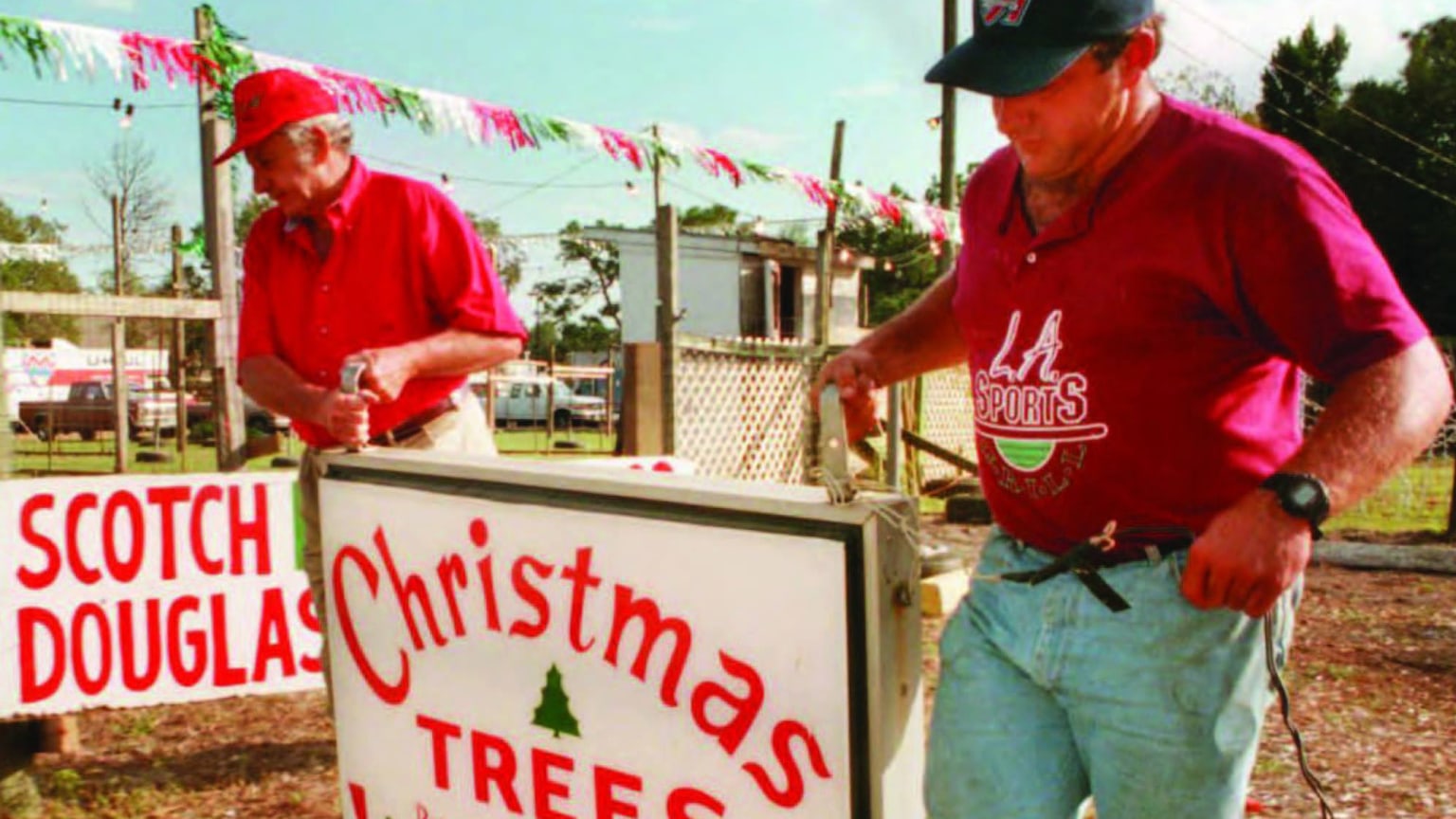 Two men in short-sleeved shirts carry a sign for Christmas trees on a sunny day