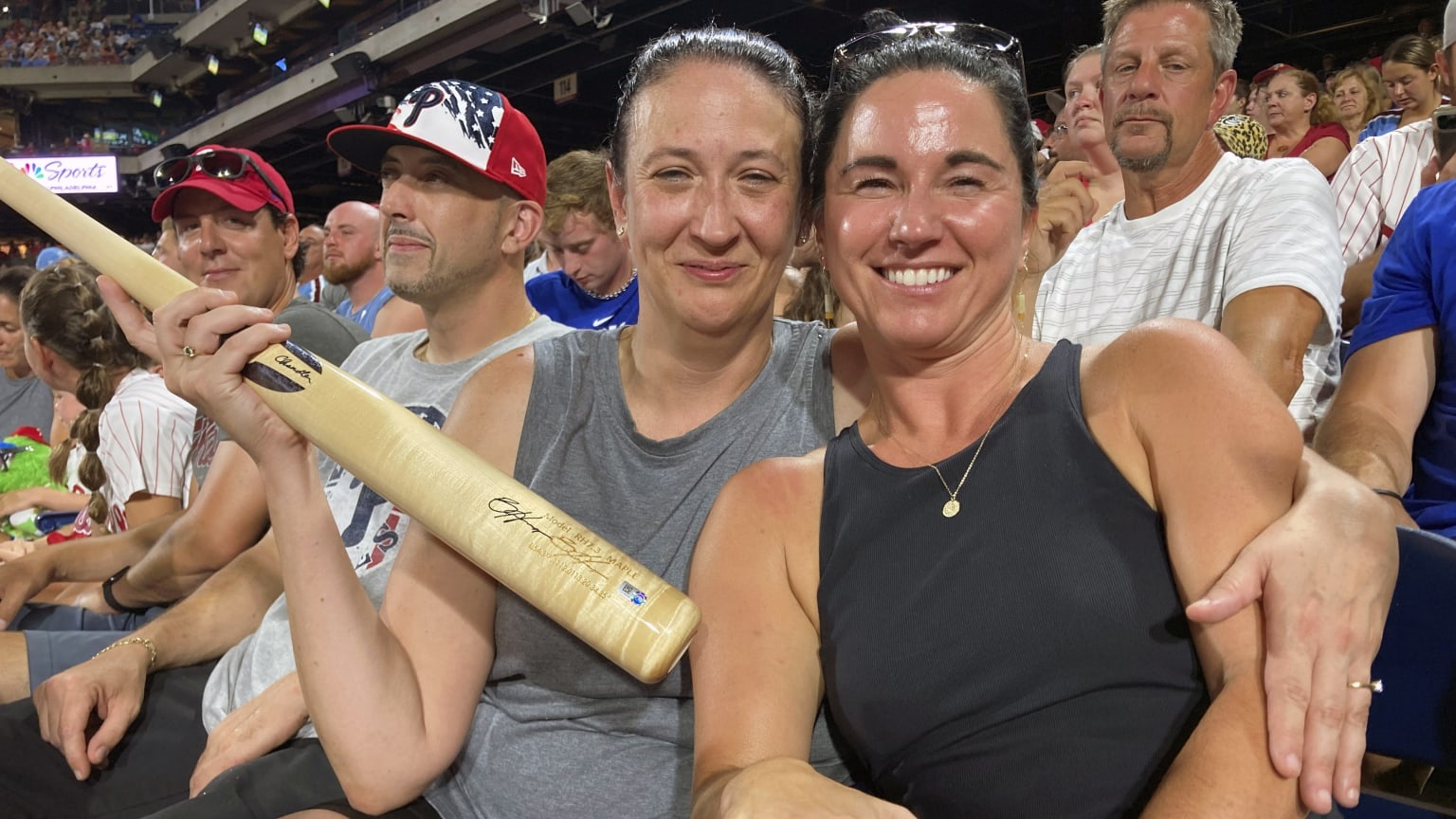 A woman at a ballgame smiles while holding a bat with a signature on it in her right hand, her left arm around another woman, who is also smiling