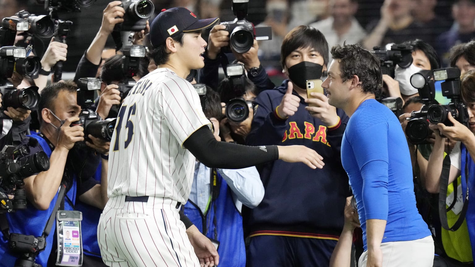Two players, one in a white uniform, one in a blue shirt, greet each other in front of a crowd of photographers