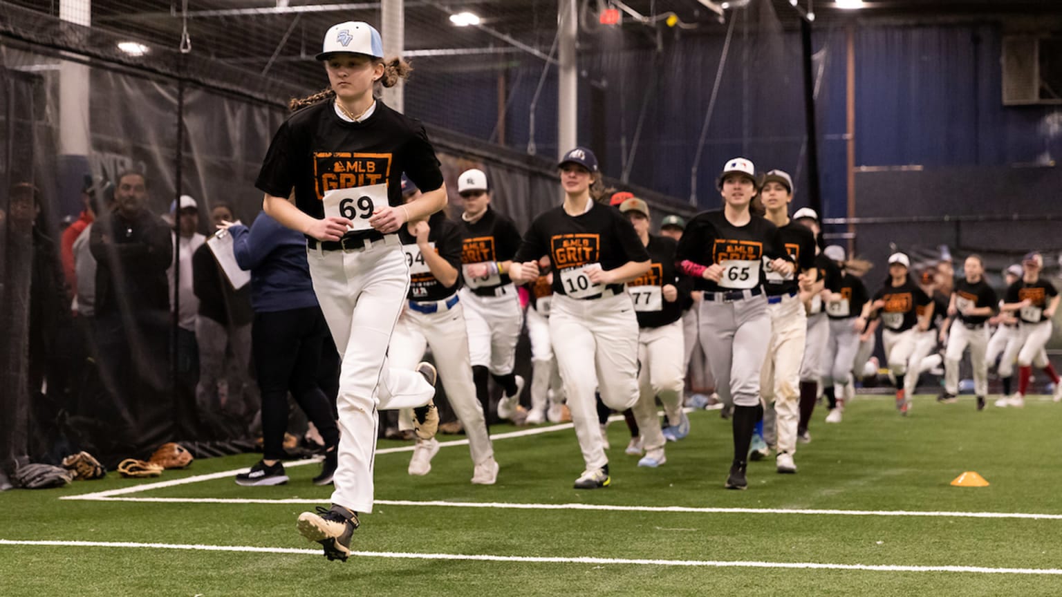 A group of girls in baseball uniforms run at an indoor facility