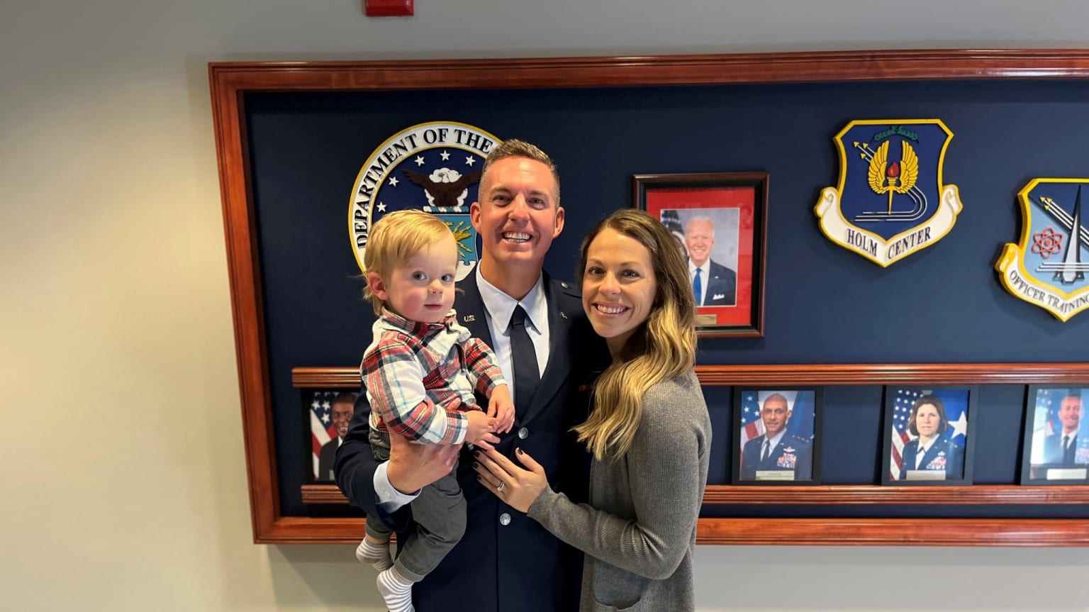 A man in an Air Force uniform smiles while holding his son, with his wife next to him. Behind them are framed photos and military logos