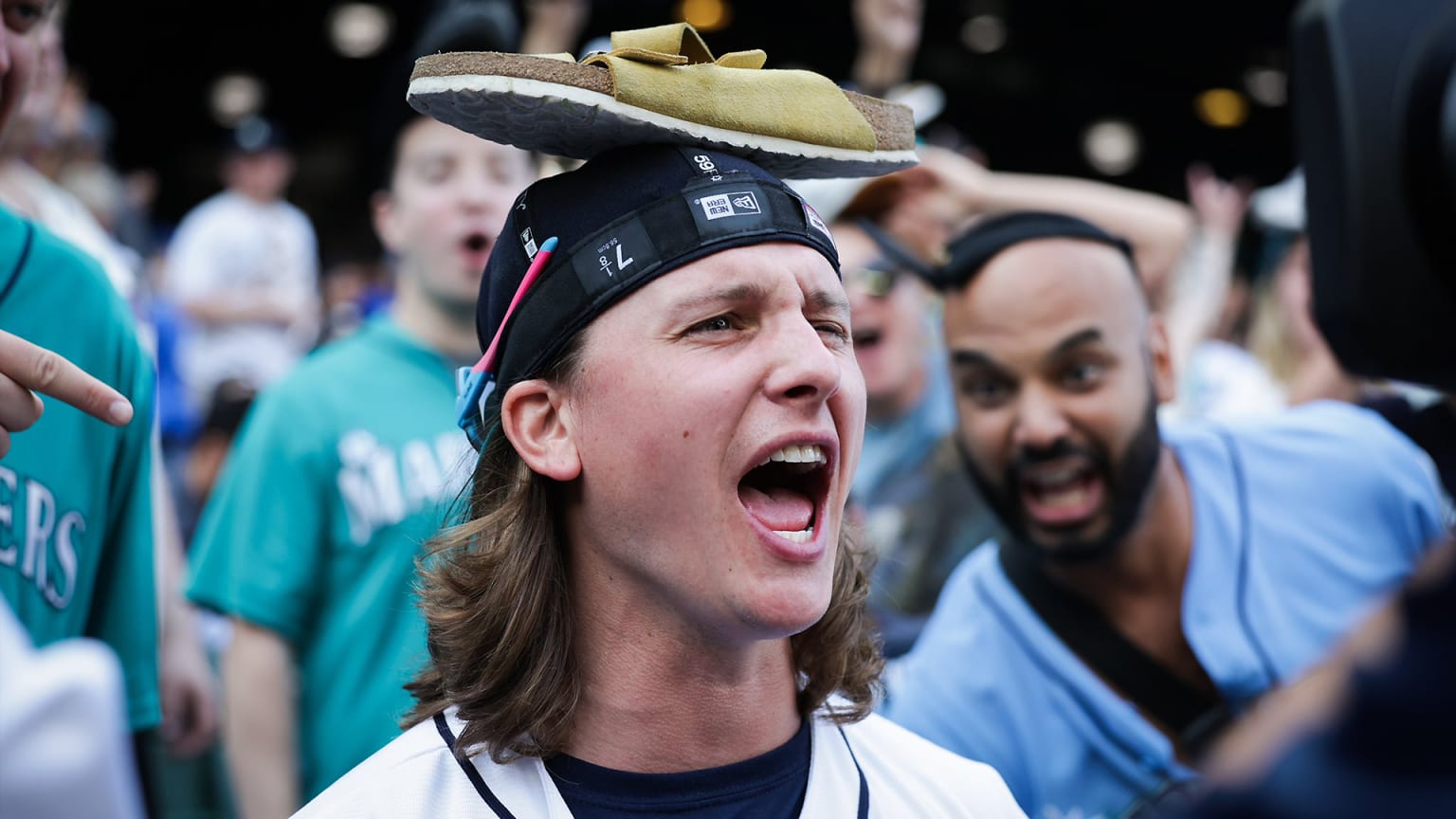 A fan with long hair cheers while wearing his cap inside-out and backwards, with a sandal balanced on top of his head