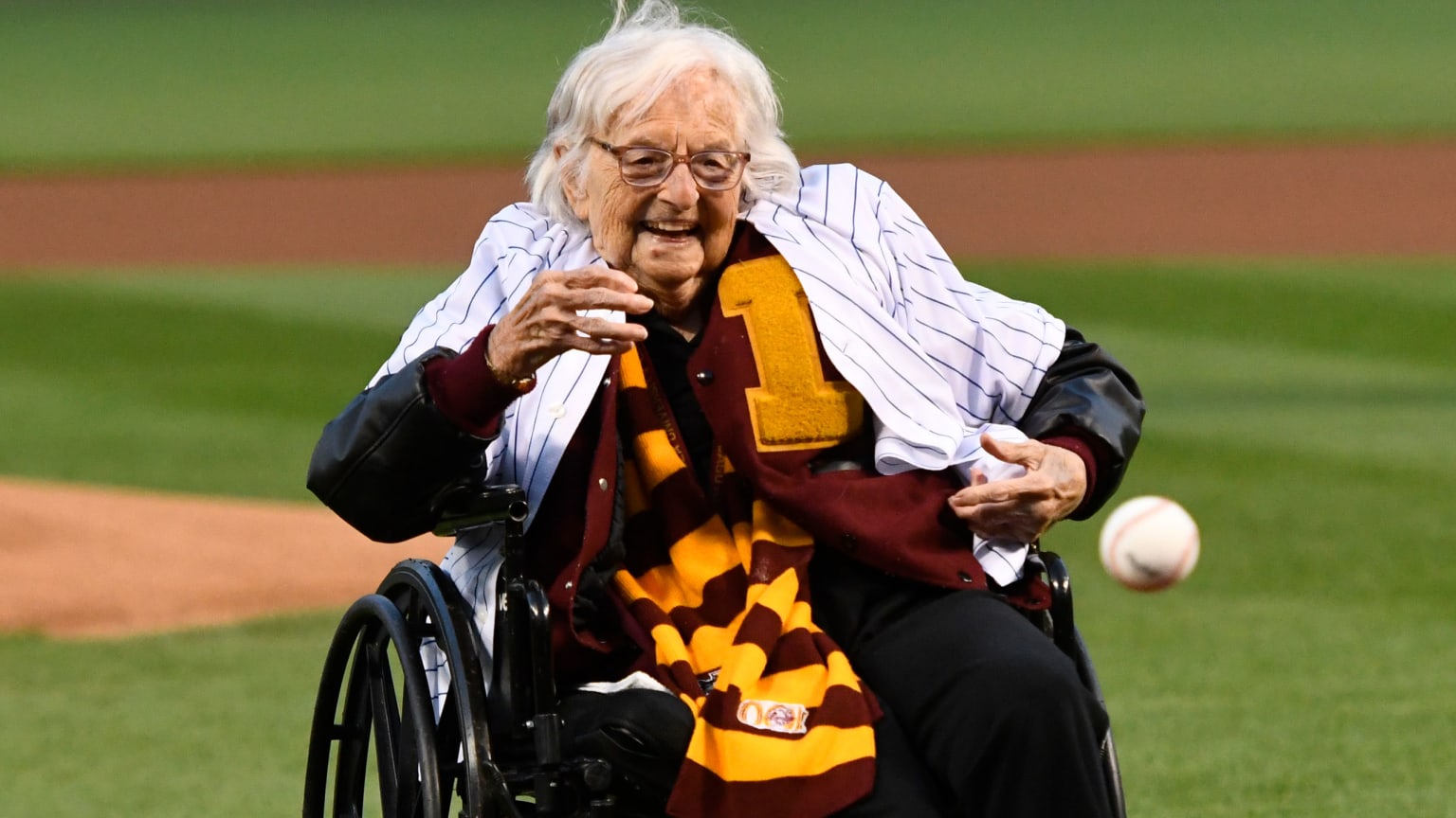 An old woman in a wheelchair wearing a white pinstriped jersey over a maroon-and-gold sweater, smiles while tossing a baseball underhand