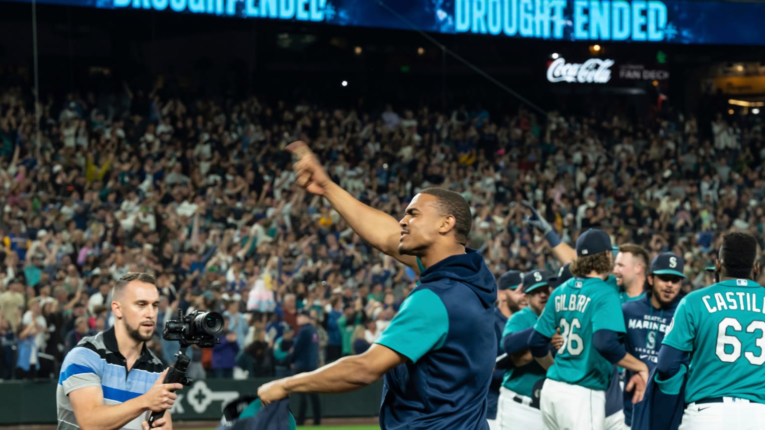 Mariners outfielder Julio Rodriguez celebrates in a sweatshirt, pointing to the fans, while a man in a polo shirt films him. Mariners players congratulate each other behind Rodriguez and to his right, all in front of a background of jubilant fans