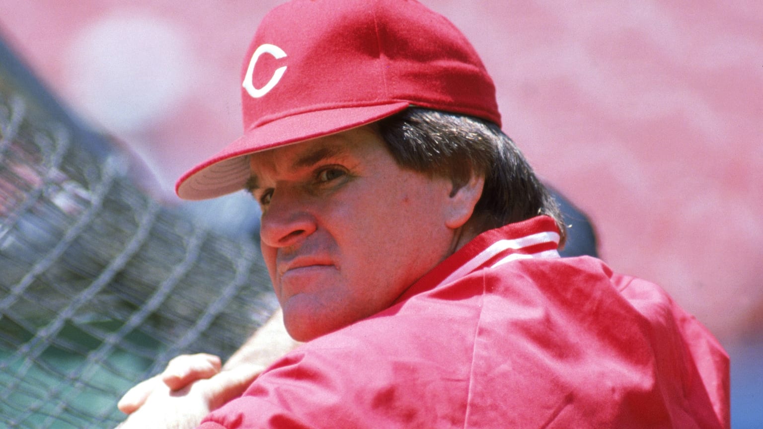 A baseball player wearing a red cap and jacket, looking serious while glancing over his shoulder against a blurred background.