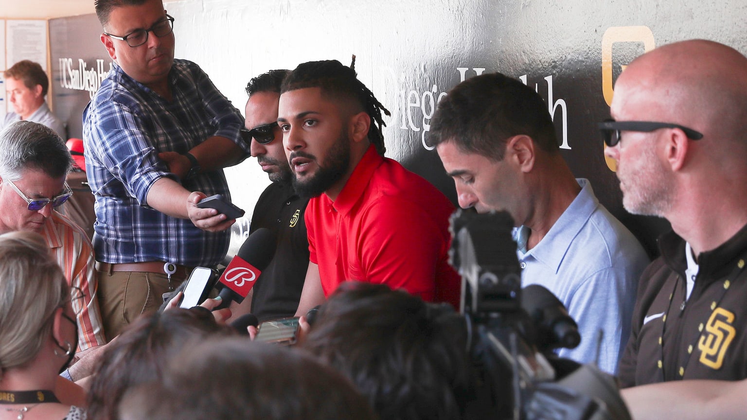 Fernando Tatis Jr., in a red shirt, sits in the dugout surrounded by reporters