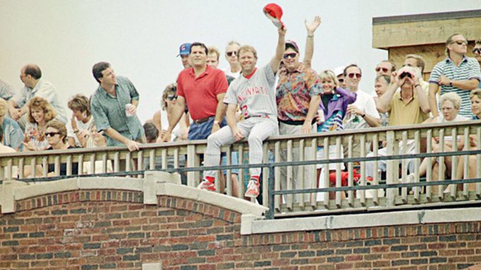 A player in a Reds uniform sits on a balcony railing and waves his cap