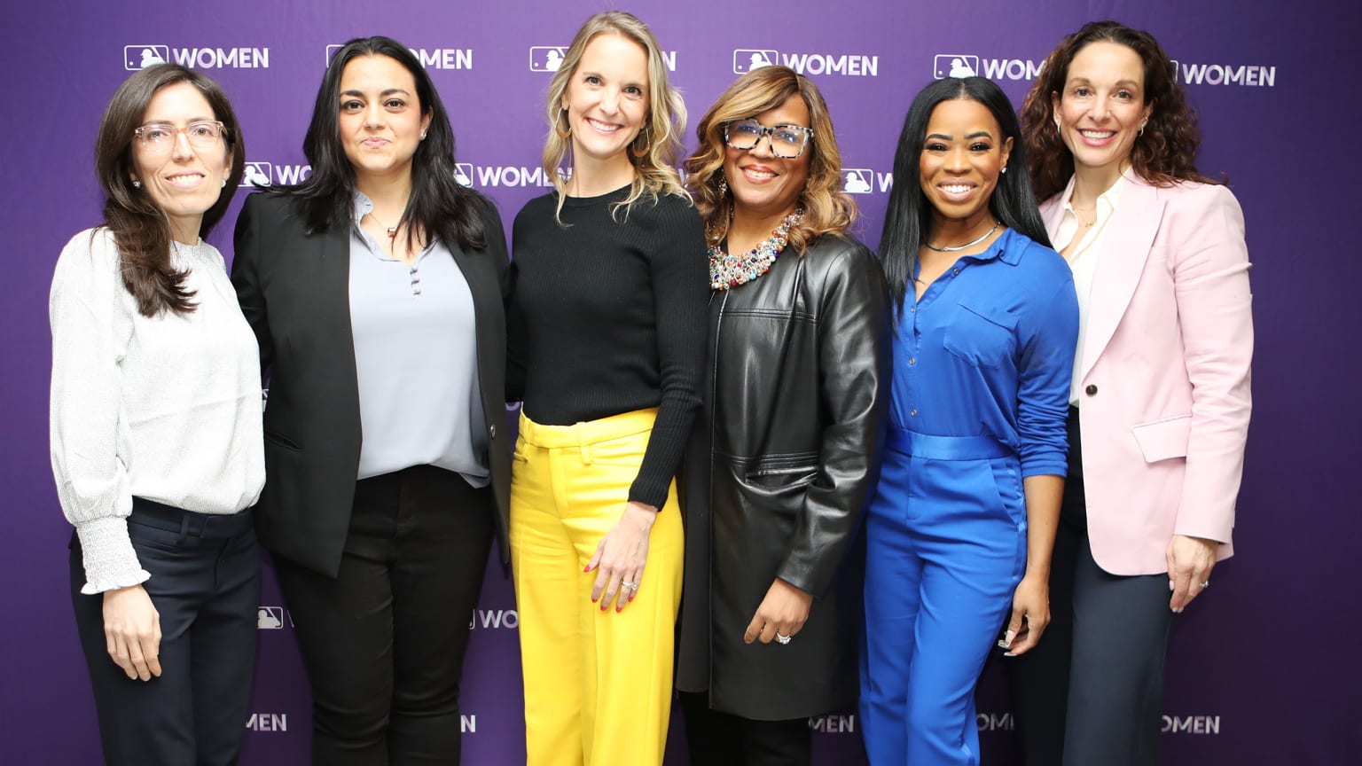 Six women stand in front of a purple backdrop for a photo
