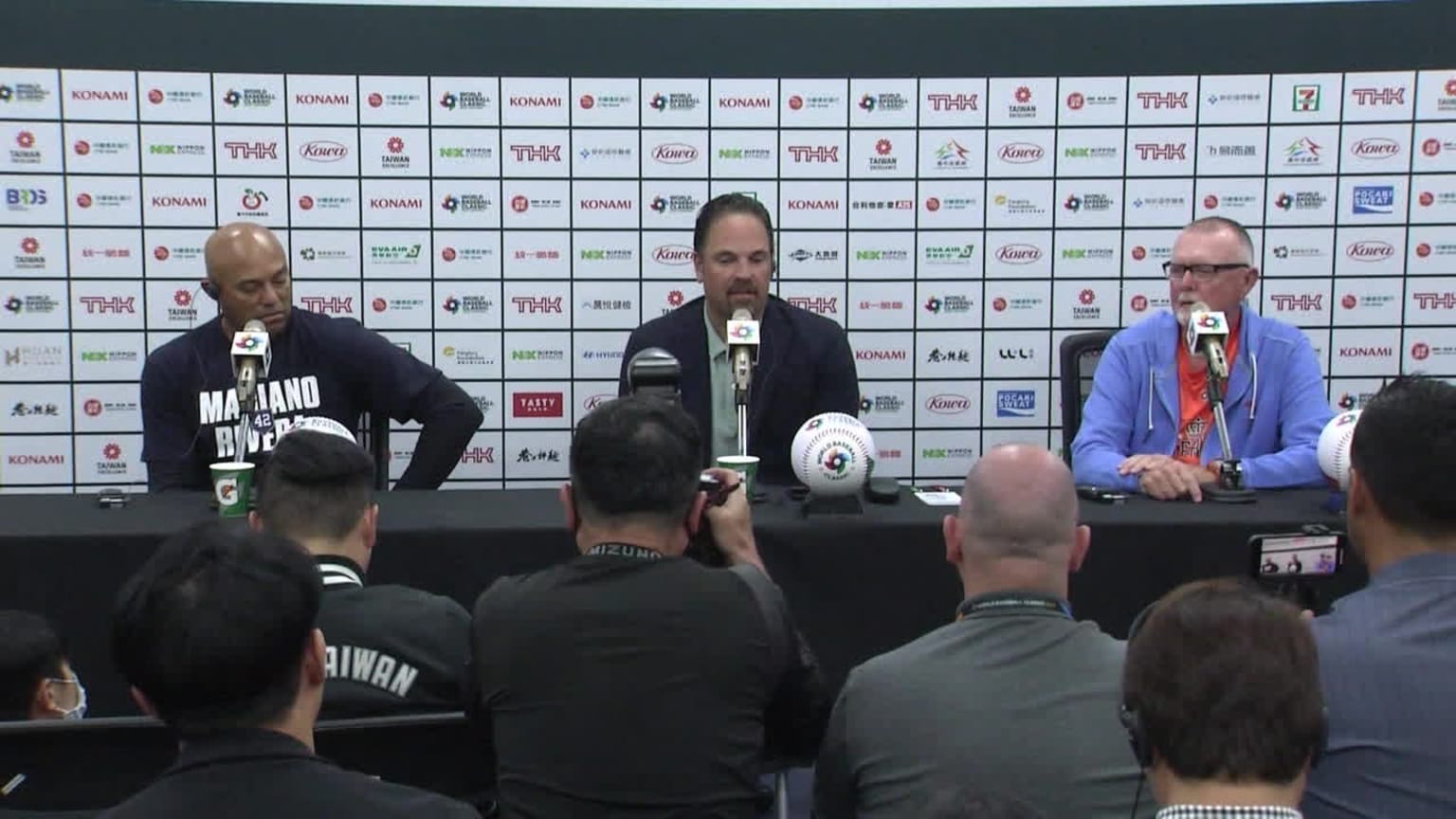 Three men sit behind microphones at a long table in front of an audience