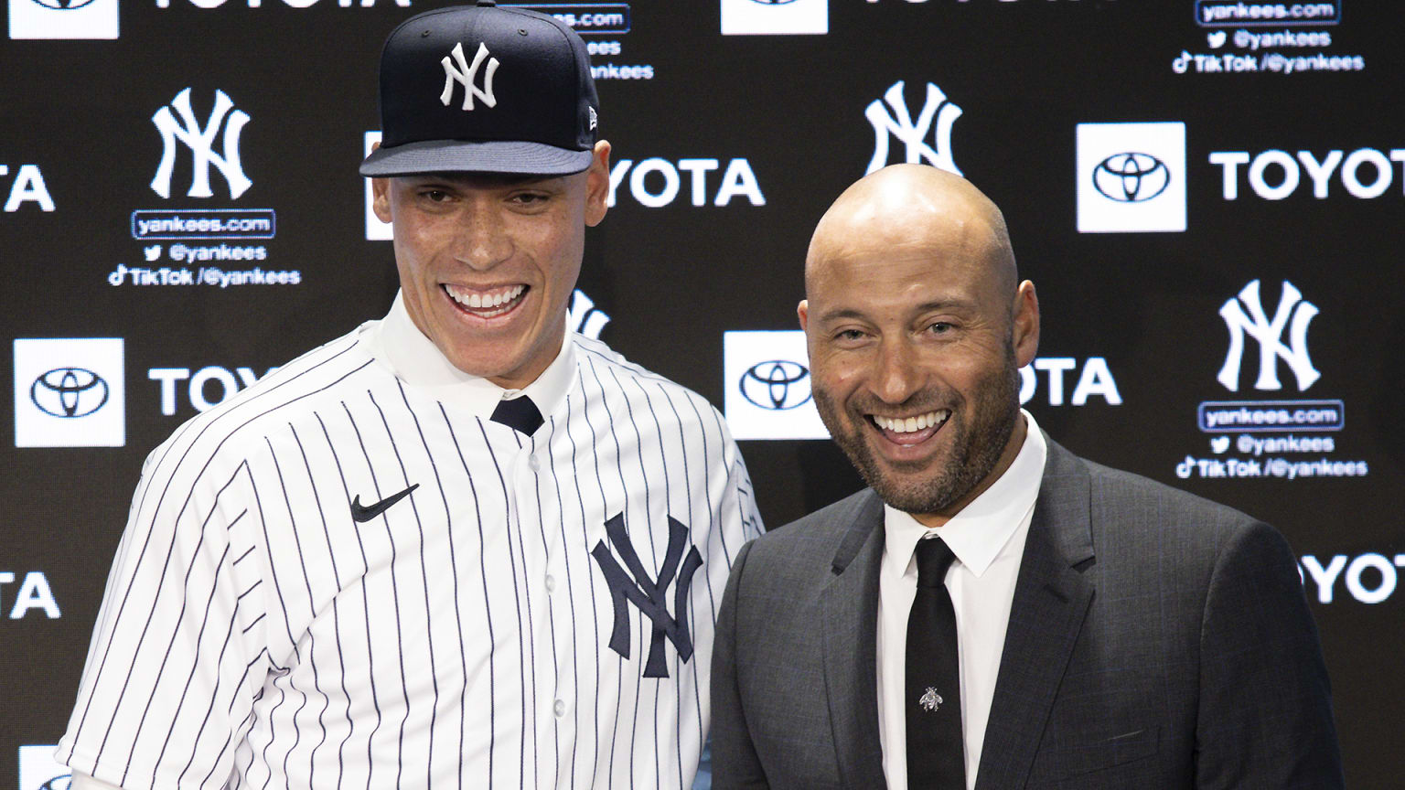 Aaron Judge in a Yankees cap and jersey smiles alongside Derek Jeter, who wears a suit