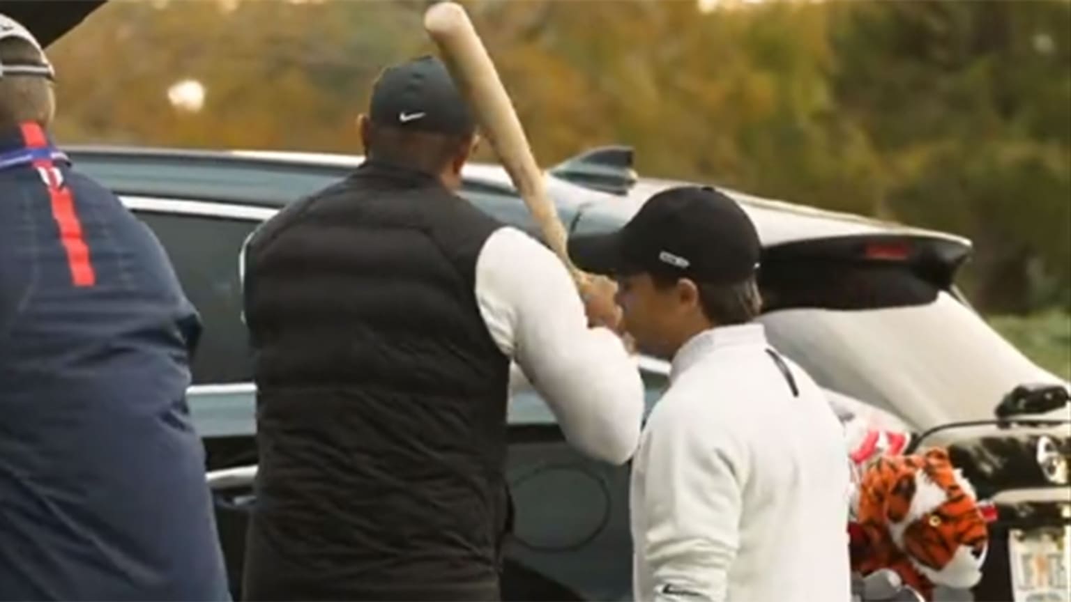 Tiger Woods, seen from behind wearing a black vest, holds a baseball bat, ready to swing