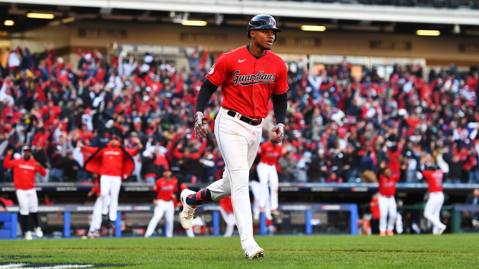 A Guardians player in a red jersey jogs to first base as teammates behind him jump out of the dugout to celebrate and fans cheer