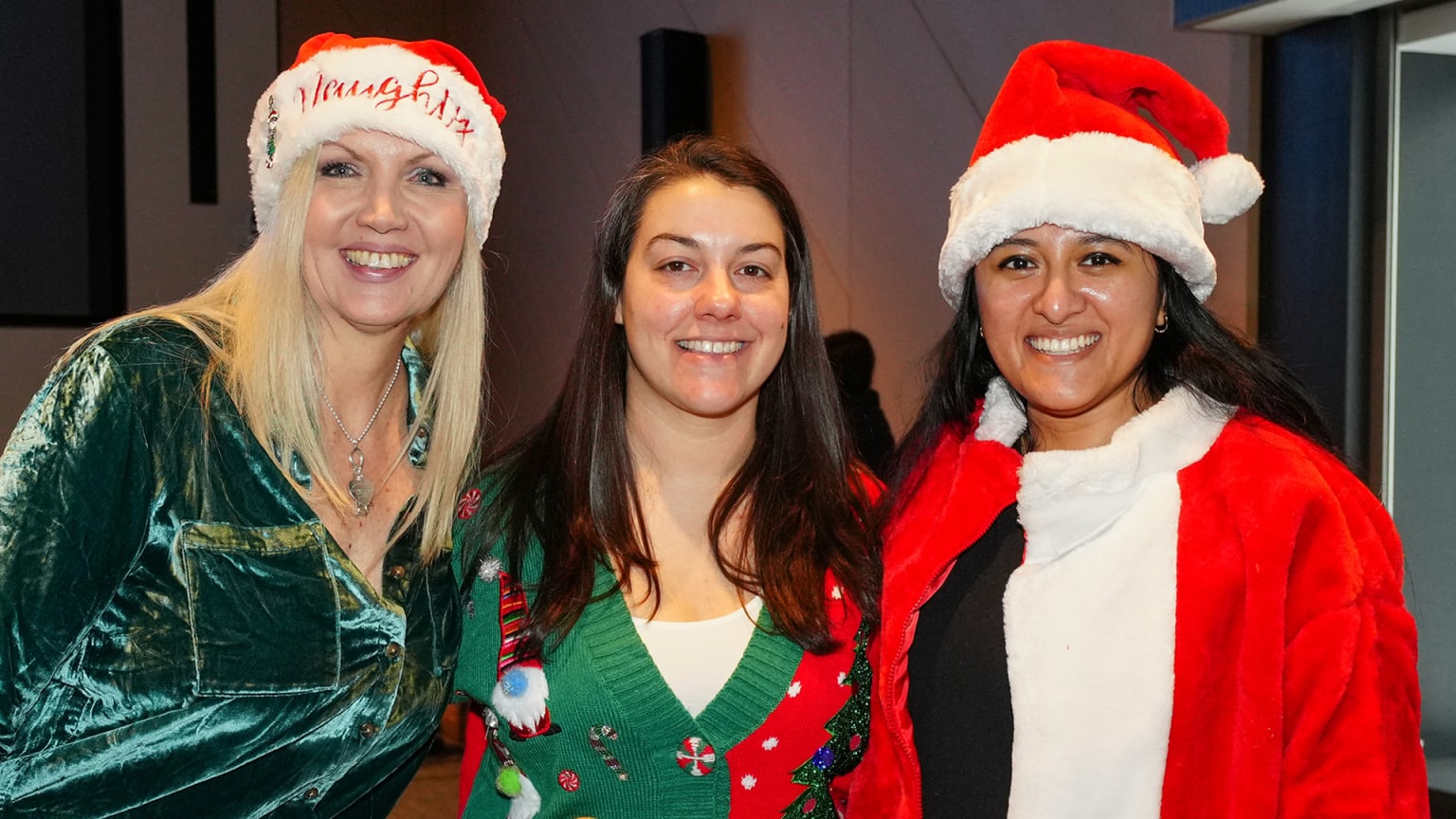3 women pose and smile in Santa hats and festive sweaters