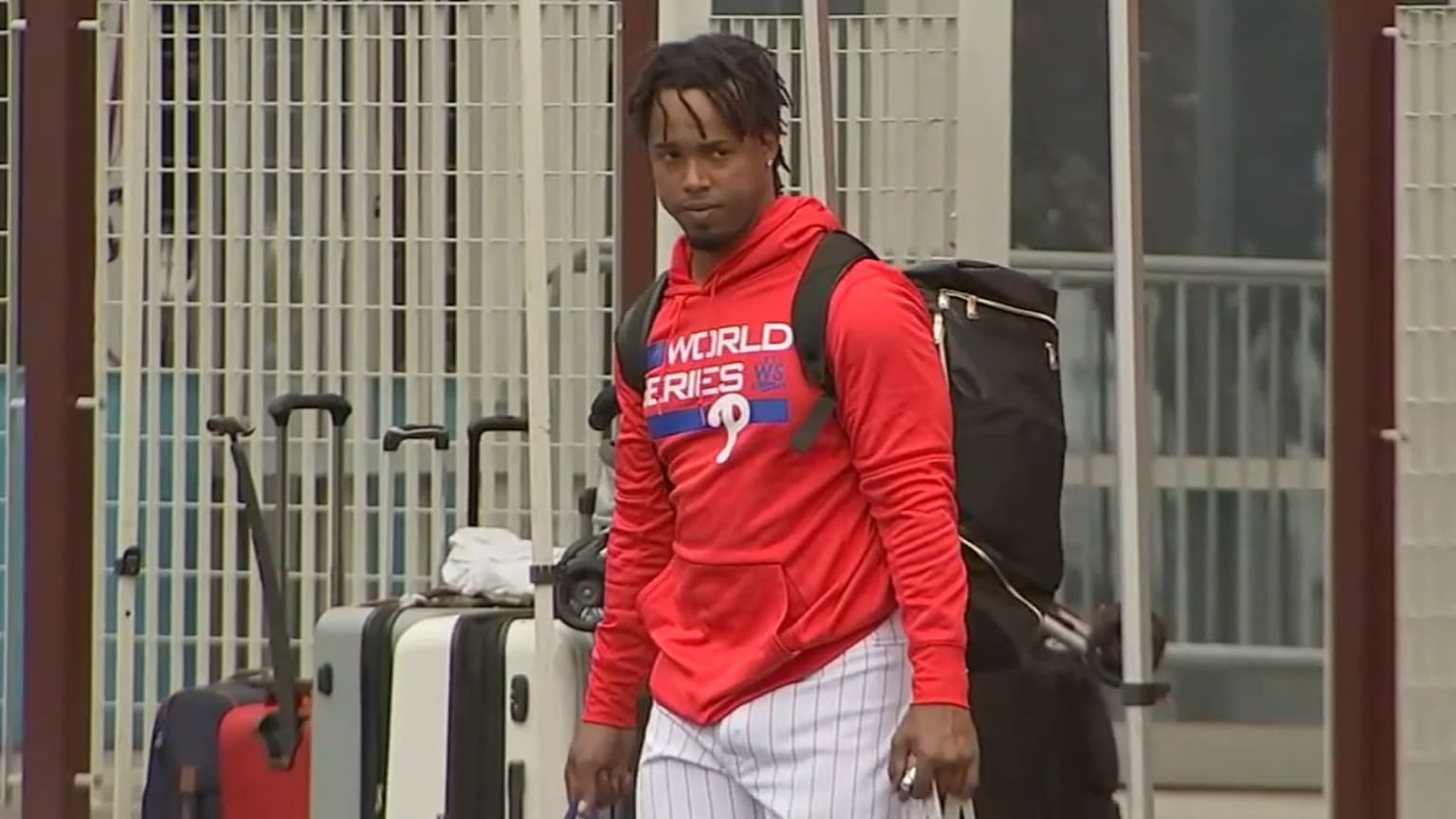 Segura stands near some luggage wearing a red hooded sweatshirt and his white pinstriped uniform pants and a backpack