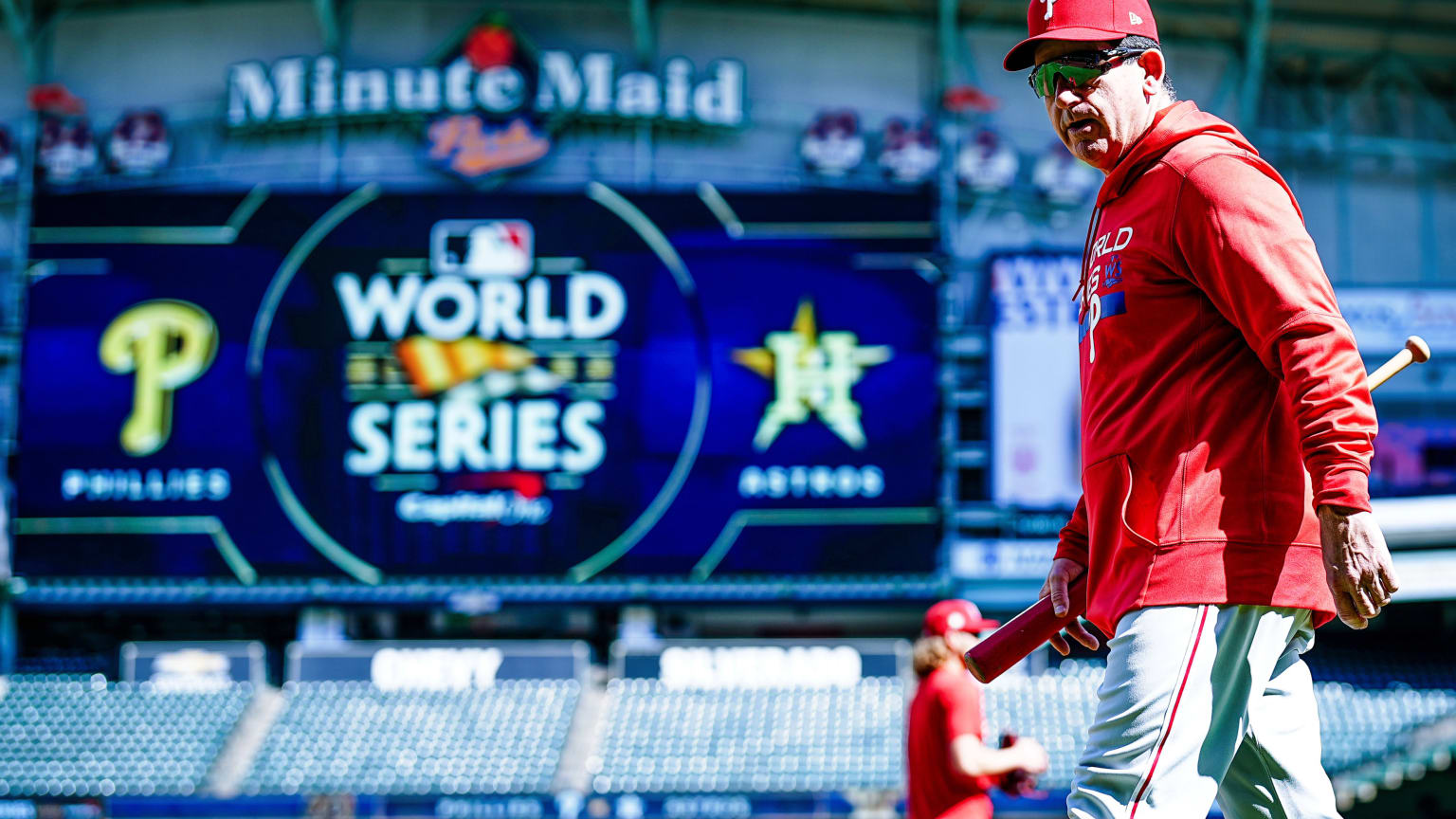 Phillies manager Rob Thomson, in a red sweatshirt and gray pants, with the scoreboard in the background showing the Phillies, World Series, and Astros logos