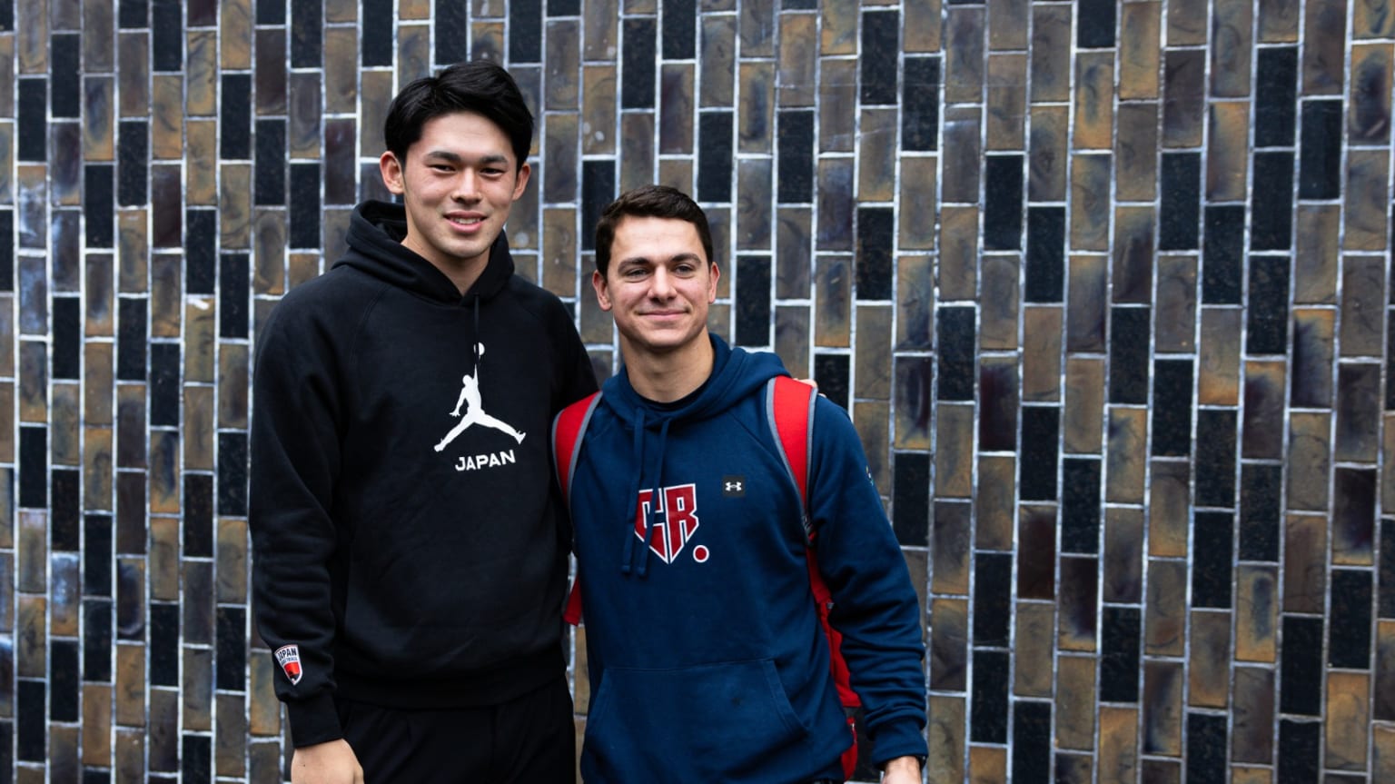 Two men in hooded sweatshirts pose together for a photo against a tiled wall