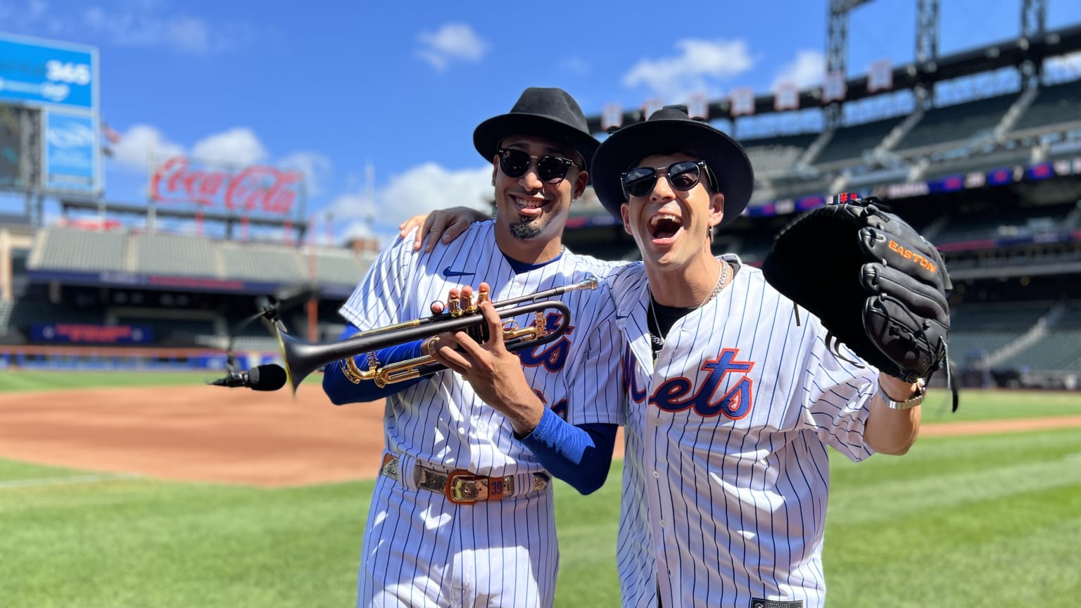 Mets closer Edwin Diaz, in his game uniform, a black fedora hat and sunglasses, holds a trumpet and smiles while musician Timmy Trumpet, in a Mets jersey, black fedora and sunglasses, drapes an arm around Diaz and smiles