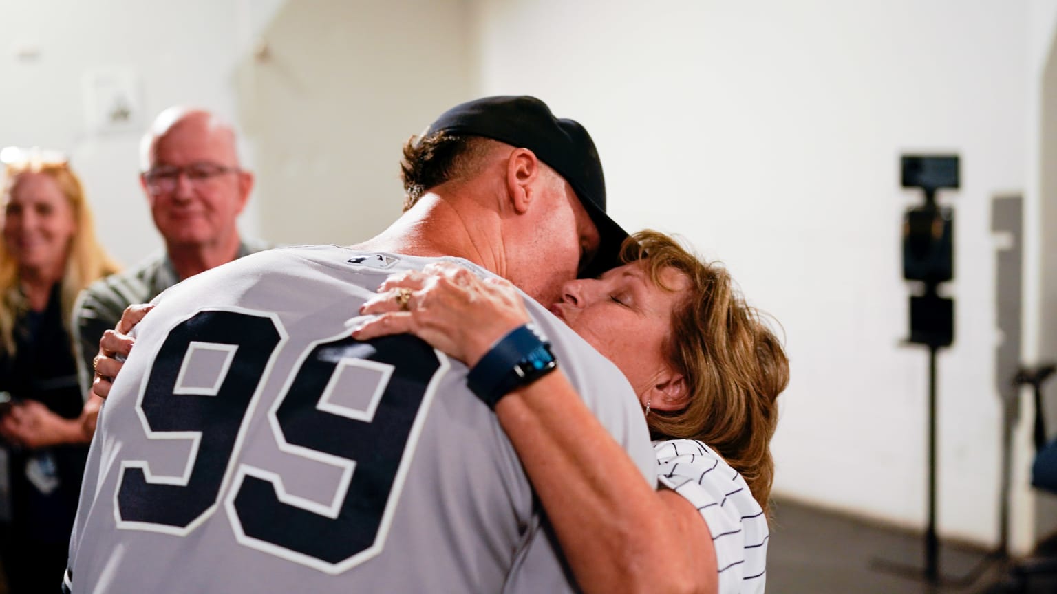 Shown from behind, the 99 on his back, Aaron Judge leans down to hug his mom and kiss her cheek