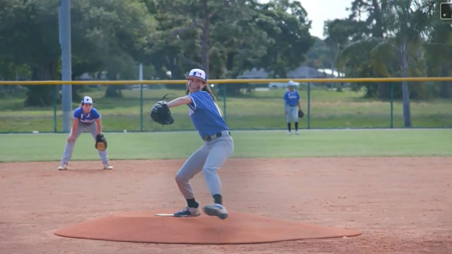 A pitcher delivers a pitch as the shortstop and center fielder watch in the background