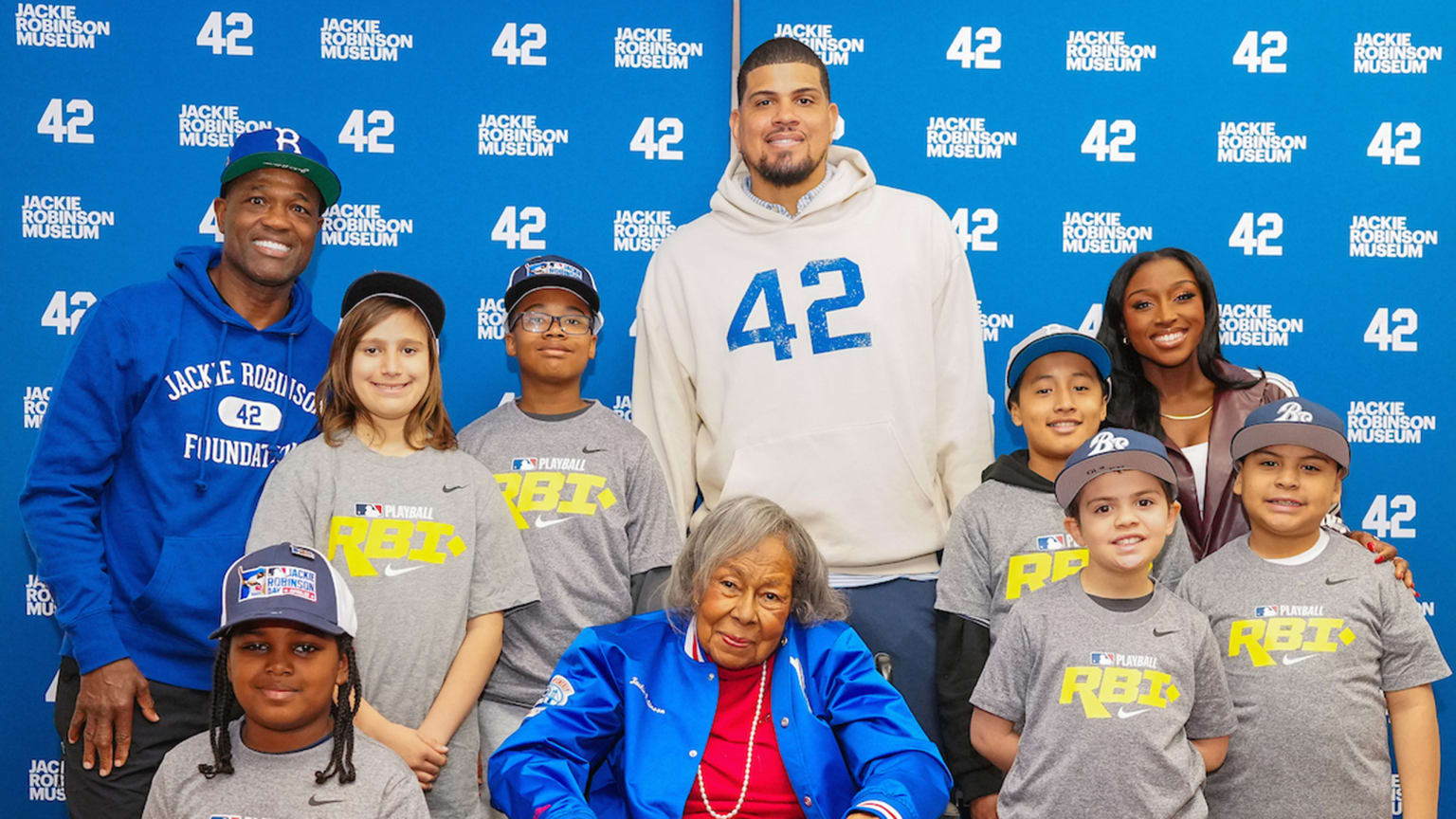 Former players Harold Reynolds and Dellin Betances, plus MLB Network host A.J. Andrews, stand with children around a seated Rachel Robinson