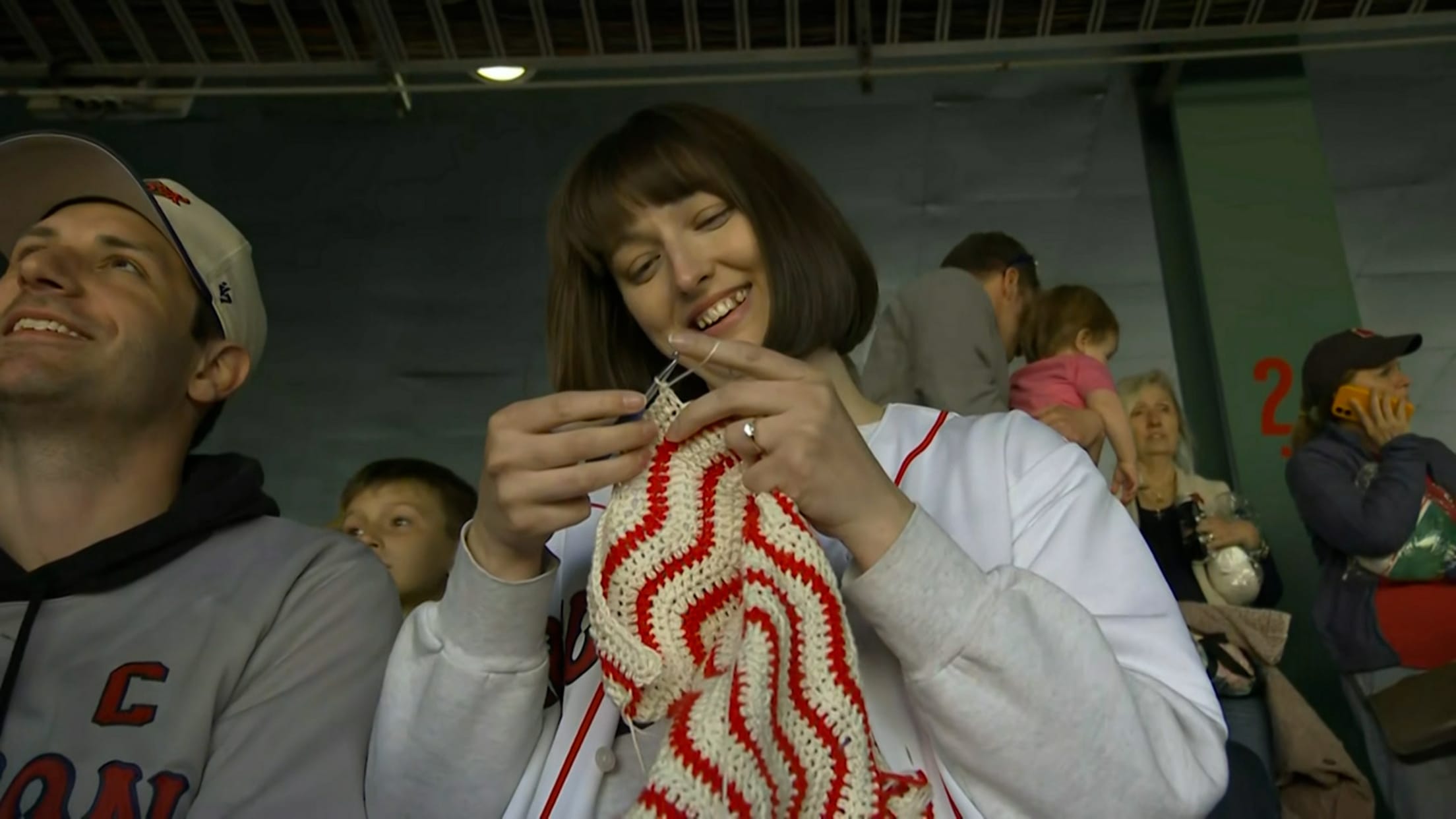 A woman in a Red Sox jersey crochets a red and white sweater at the ballpark