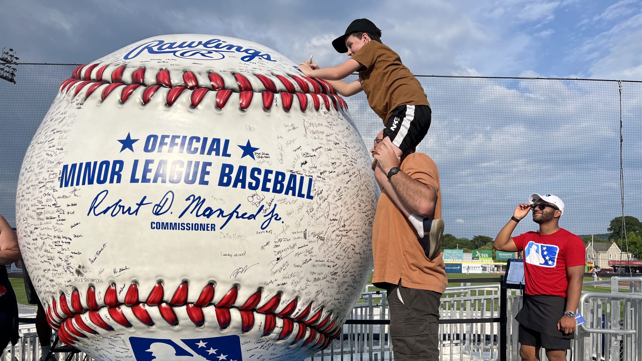 A child on a person's shoulders signs a huge replica baseball