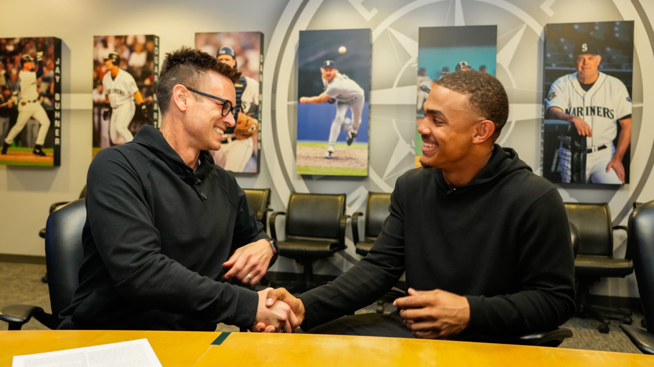 Mariners GM Jerry Dipoto and outfielder Julio Rodriguez smile and shake hands at a conference table in a room with photos of Mariners players on the wall