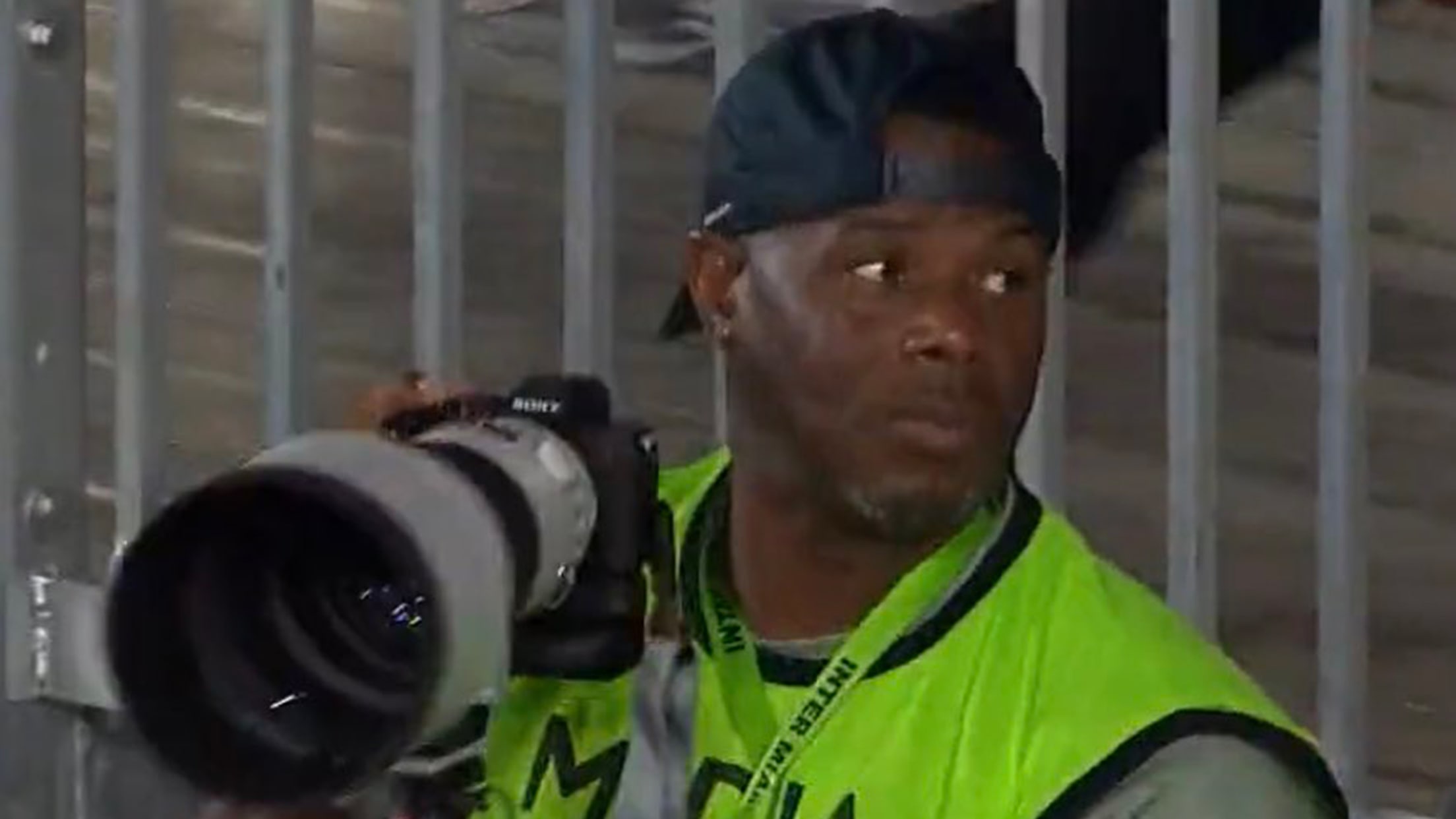 Ken Griffey Jr., wearing a bright yellow-green vest, holds a camera with a long telephoto lens