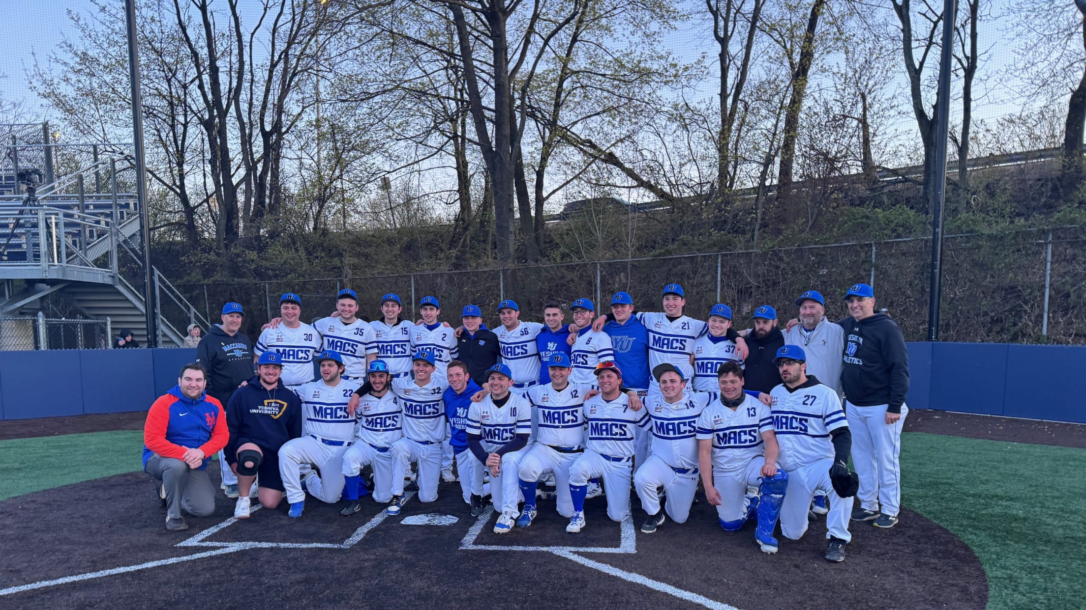 The Yeshiva University Maccabees baseball team poses at home plate on a suburban baseball field with a steep slope and trees behind the backstop