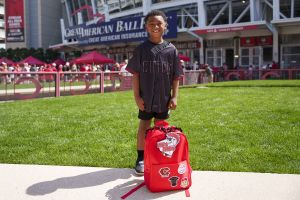  Happy Reds Heads member standing behind their brand new bright red Reds Heads backpack with patches of a black Reds jersey, a baseball and a Reds City Connect logo. 