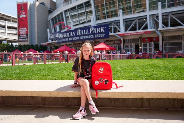  Proud Reds Heads member seated next to their backpack outside Great American Ballpark. 