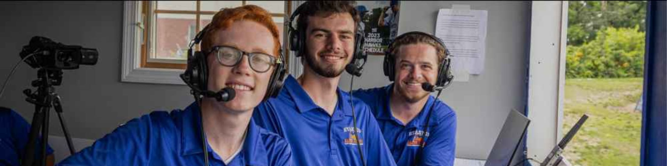 Three Hyannis broadcasting interns sit in the press area, smiling for the camera in-between broadcasting the game.